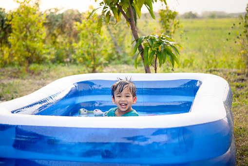 Portrait of asian child, kid or little boy to smile, bath, play water or funny activity in portable inflatable swimming pool at outdoor, backyard or garden in summer evening. Include green field, sky background.