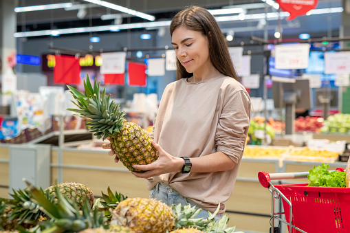 Young woman wearing casual outfit choosing best pinapple in modern supermarket, copy space