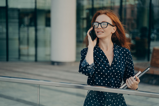 Thoughtful redhead woman wears spectacles polka dot dress has telephone conversation looks into distance stands near blurred office background carries necessary things for working. Lifestyle