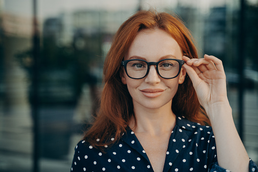 Portrait of confident beautiful red-haired business woman adjusting spectacles eyeglasses while posing on city street in summer, looking at camera with confidence, blurred urban background