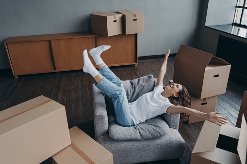Overjoyed european woman has bought a new house. Spanish young lady relaxing in armchair among boxes. Concept of opportunity for woman and mortgage. Independence and new lifestyle.