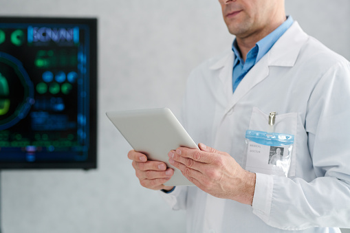 Close-up of mature doctor in white coat using digital tablet for online work
