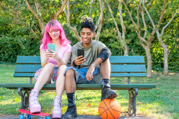 alternative diverse couple hanging out together sitting in a park bench holding smartphone online on social media web. interracial teenagers friends relaxing outdoors. tech and lifestyle concept pareja diversa alternativa pasando el rato juntos sentados en un banco del parque sosteniendo un teléfono inteligente en línea en la web de las redes sociales. adolescentes interraciales amigos relajándose al aire libre. concepto de tecnología y estilo