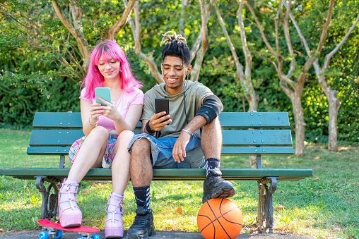 alternative diverse couple hanging out together sitting in a park bench holding smartphone online on social media web. interracial teenagers friends relaxing outdoors. tech and lifestyle concept