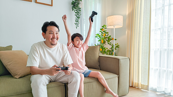 Asian father playing video games with his young boy at home, competing and enjoying themselves, smiling