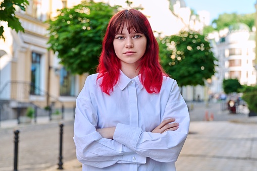 Serious confident young female with crossed arms looking at camera. Teenage beautiful fashionable girl with red dyed hair in city. Beauty, youth, lifestyle concept