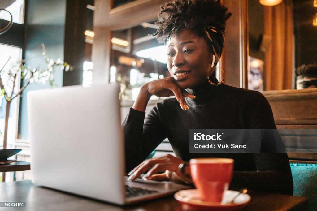 African woman on a online meeting in a cafe A young African woman having a work meeting call in a cafe. Young Adult Stock Photo