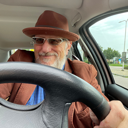 Smiling senior man with glasses, grey beard, brown jacket and hat drives a car