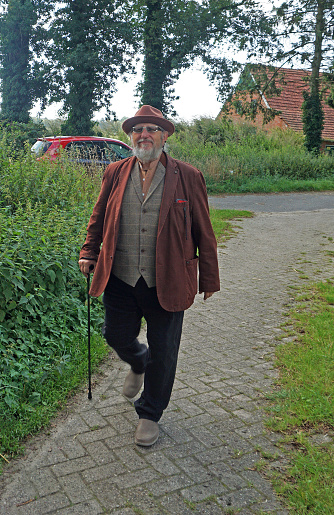 A senior male hiker with a stick. A brown jacket and a brown hat in a rural environment
