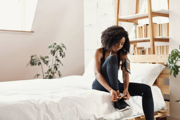 One fit, focused young female athlete tying her laces while getting ready to go on a run. A young African woman listening to music and getting ready for a workout in her bedroom at home
