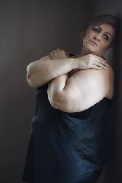 mujer gorda solitaria y deprimida de pie y mirando por la ventana de casa. - mature adult sadness overweight women fotografías e imágenes de stock