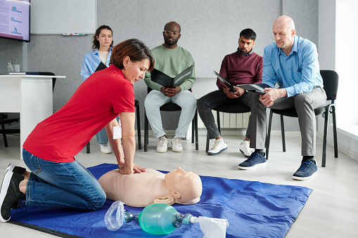 Medical instructor showing CPR on training mannequin on floor to people who sitting on chairs and watching during lesson