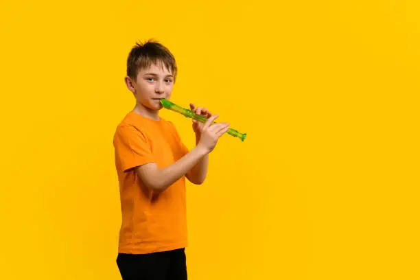 Boy 9-11 y.o in Basic Orange T-shirt Plays Block Flute and Looks at Camera on Yellow Background. Concept of Music Education and Training in Music School on Wind Instruments