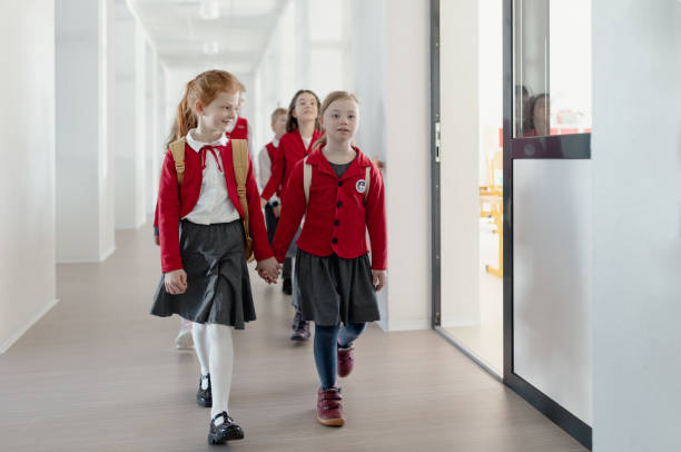 istock Happy schoolgrirl with Down syndrome in uniform holding hands with her classmate walking in scool corridor with classmates behind them. 1401147979