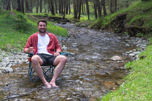 Man sitting in his camping chair in the stream running through the forest. The man sitting in the camp chair in the stream has a glass in his hand. Shot with a full-frame camera in daylight.