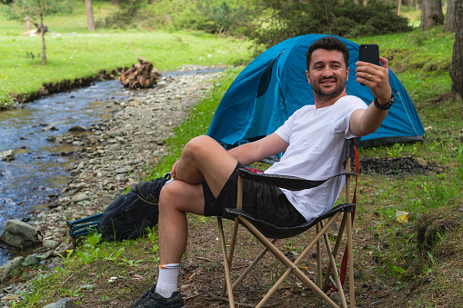 The man camping by the stream passing through the forest is taking selfies with his mobile phone. A camping tent was set up on the flat meadow in the forest. The image of the stream flowing next to the tent was taken with a full frame camera.