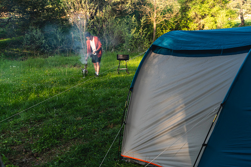 man camping in the forest is cooking on the barbecue. He set up his tent under the tree. Shot with a full-frame camera in daylight on a grassy field.