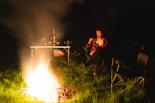 Image of man camping in forest at dusk. The man who lights the campfire is warming up and drinking tea. Shot with a full frame camera.