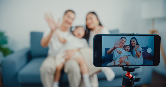Young Asian parents sitting on couch use smartphone say hello with camera in living room at home.