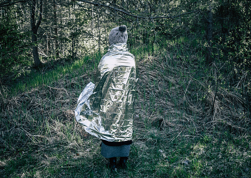 Woman person is lost and stand in wild forest in cold day, using first aid emergency blanket to prevent hypothermia and body heat loss. Emergency blanket concept.