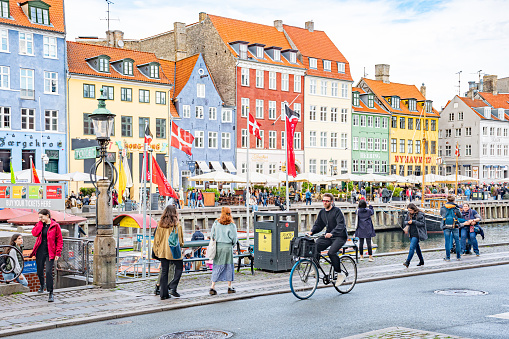 Copenhagen, Denmark - May 23 2022: Cyclist, pedestrians and tourists in Christianshavn neighborhood, Copenhagen, Denmark with traditional Danish colorful houses, boats and flags on background