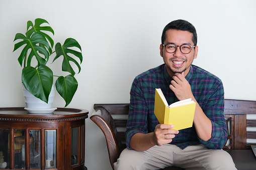 Adult Asian man sitting in a couch while holding a book and showing happy expression