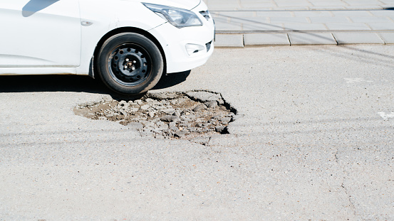 Side view of a car driving on a broken road with a hole, dangerous driving on damaged asphalt outdoors, close-up. Selective focus on pit in road surface.