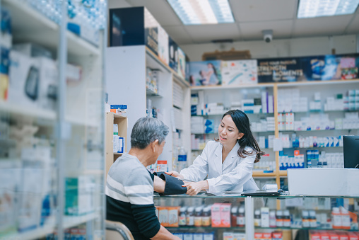 asian chinese female pharmacist measuring blood pressure with senior man at drug store pharmacy