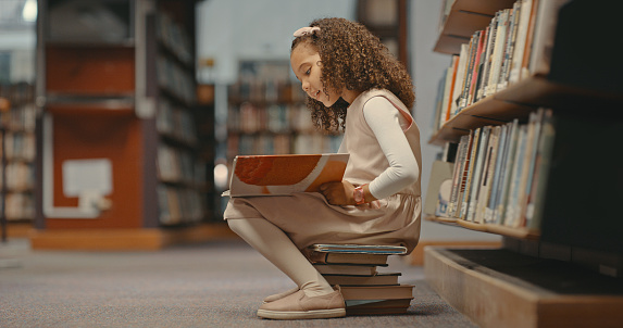 Young girl sitting on books in the library and reading a book. Cute girl with curly hair doing her project. Female alone and doing research for a project