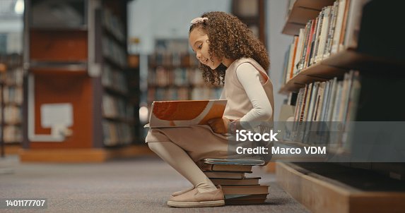 istock Young girl sitting on books in the library and reading a book. Cute girl with curly hair doing her project. Female alone and doing research for a project 1401177707