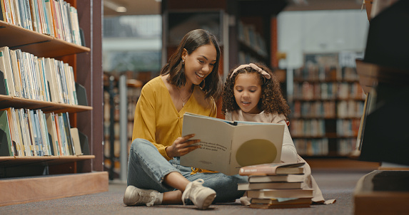 Two females on the floor reading a book in the library. Young mother helping her daughter looking at a book together. Woman and girl doing a project