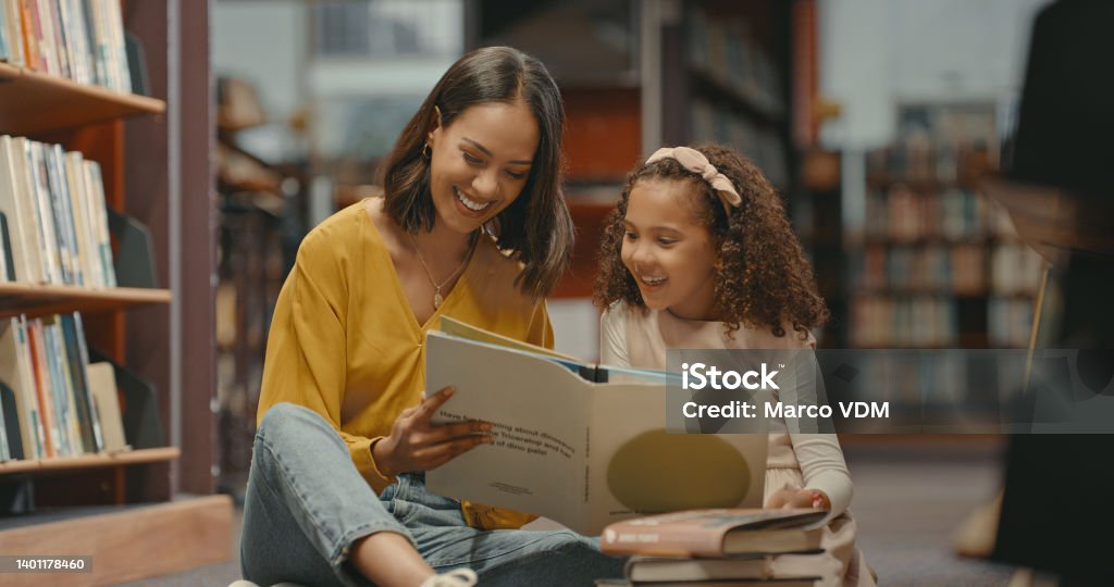 Teacher helping a young student with her homework in the library after school. Two females are reading a book together in the bookstore. They are doing research for a project Child Stock Photo