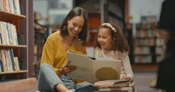 Photo of Teacher helping a young student with her homework in the library after school. Two females are reading a book together in the bookstore. They are doing research for a project