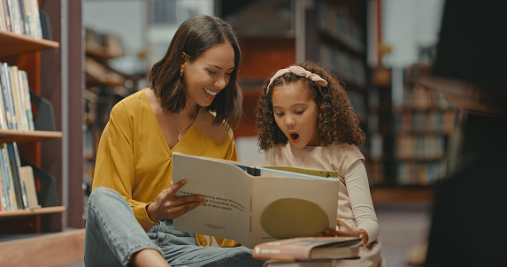 Mother and daughter reading a book together in the library. Cute daughter doing her homework at the library with her mother. Two mixed race females inside the library