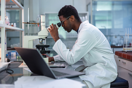 Portrait of African-American male scientist wearing a lab coat, looking under a microscope, analyzing a sample during his work in a laboratory.