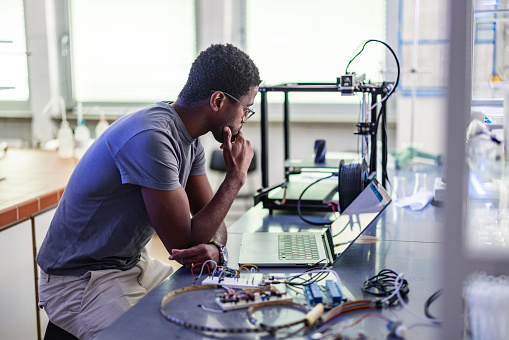 Young African-American technician sitting in the chair, thinking and repairing his 3D printer in the laboratory.