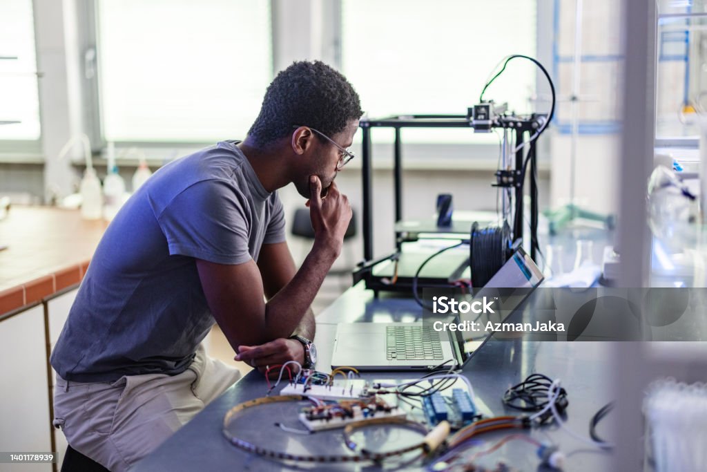 Young Designer During His Work In Laboratory Young African-American technician sitting in the chair, thinking and repairing his 3D printer in the laboratory. 3D Printing Stock Photo