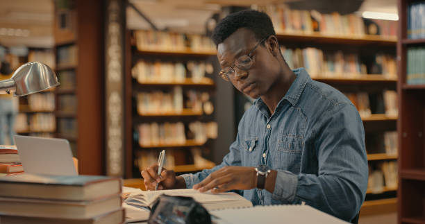 図書館の中に一人で座って研究をしているアフリカ人男性。プロジェクトに取り組んでいる人。事件の調査をしている青年。事件に取り組んでいる弁護士 - 文学 ストックフォトと画像