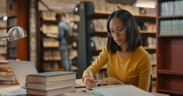 kobieta mieszanej rasy pisząca w bibliotece. młoda dama pracująca nad projektem sama. kobieta siedząca przed książkami prowadząca badania - male education college student nerd zdjęcia i obrazy z banku zdjęć