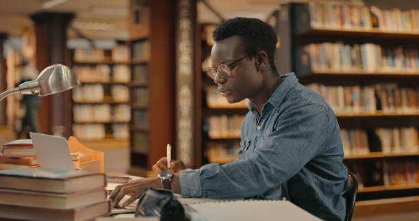 istock Male student sitting inside a library working on a project. Student sitting and doing research. African American male working on case 1401178953