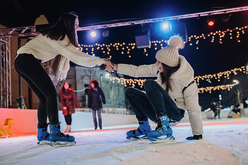 Beautiful smiling young woman helping female friend to get up while ice-skating on outdoor skate rink at night