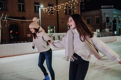 Smiling beautiful young women holding each other's hands while ice-skating on outdoor skate rink at night