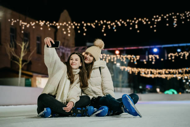 Young women taking selfie together with mobile phone while sitting on skate rink at night stock photo