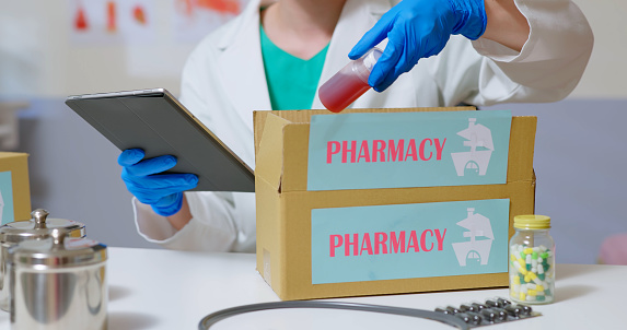 close up side view of asian male doctor wear white lab coat and surgical glove taking out checking medication with tablet from pharmacy delivery service package box in office