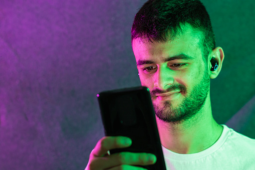 Portrait of a young bearded Caucasian man using his cell phone while wearing a wireless headset. It is illuminated with green and magenta colored lights.
