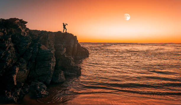 silhouette de saxophoniste jouant sur la falaise au coucher du soleil et pleine lune en arrière-plan - water wave sea tranquil scene photos et images de collection