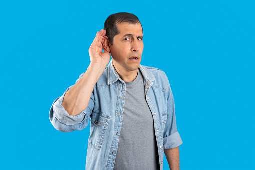 Studio shot of a handsome  man with over isolated blue background smiling with hand over ear listening and hearing to rumor or gossip. Deafness concept.