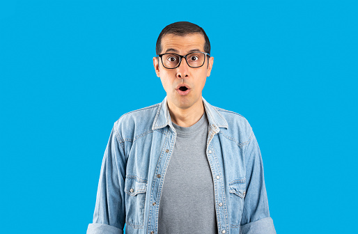 Studio shot of a hispanic man wearing glasses over isolated blue background afraid and shocked with surprise expression, fear and excited face.