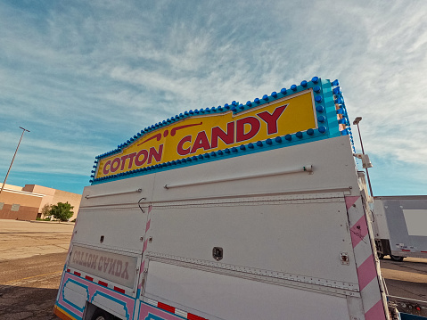 Traveling carnival lands at a local mall. Large cotton candy booth with lit sign. Sign says, \