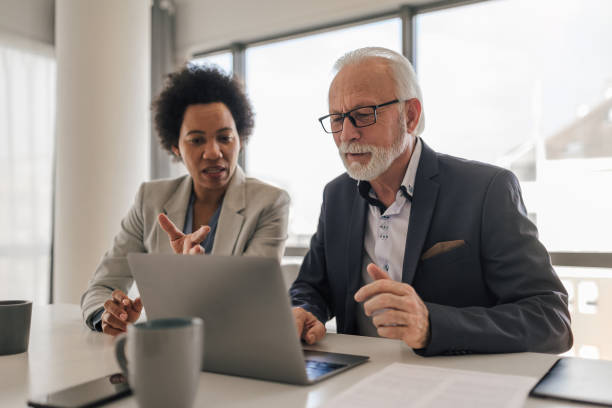 Executive discussing with senior professional while working on laptop at office Female executive discussing with senior professional while working on laptop at desk in office mature adult stock pictures, royalty-free photos & images